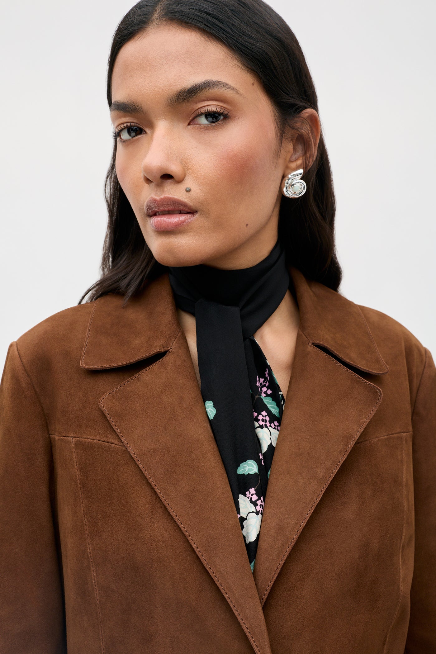 Wearing the Lainey - Chestnut brown suede jacket, a woman with long dark hair pairs it with a black floral top, black neck scarf, and large silver earrings, gazing confidently at the camera against a plain light background.