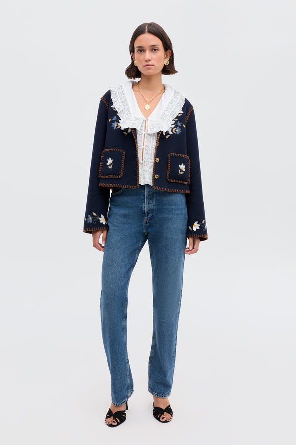 A woman faces forward in the Sunday - Petal Embroidery Navy, a navy wool-blend jacket with floral embroidery, styled with a white lace-collared blouse, blue jeans, black heeled sandals, layered gold necklaces, and a plain light background.