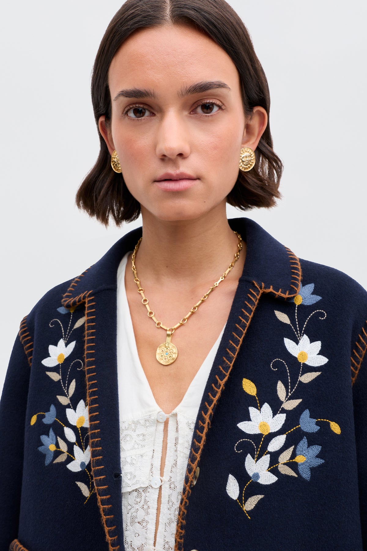 A woman with short brown hair wears gold earrings and a gold necklace. She is dressed in the Sunday - Petal Embroidery Navy jacket, a luxurious navy wool blend with white, blue, and yellow floral embroidery, over a lace-detailed white top.