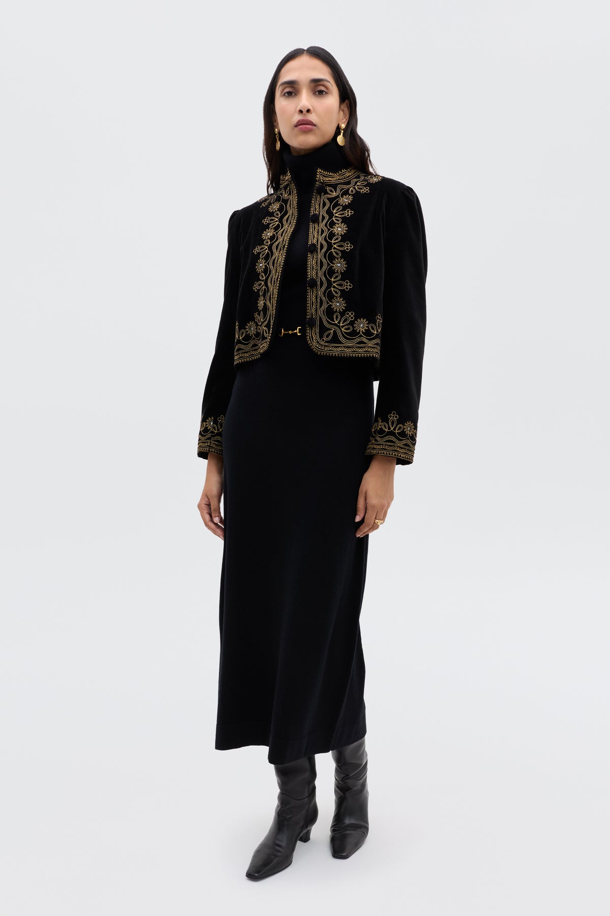 A woman poses against a plain light background in the Denver - Soutache Black: a long black dress with a cropped velvet jacket adorned in gold embroidery, styled with gold hoop earrings and black heeled boots.