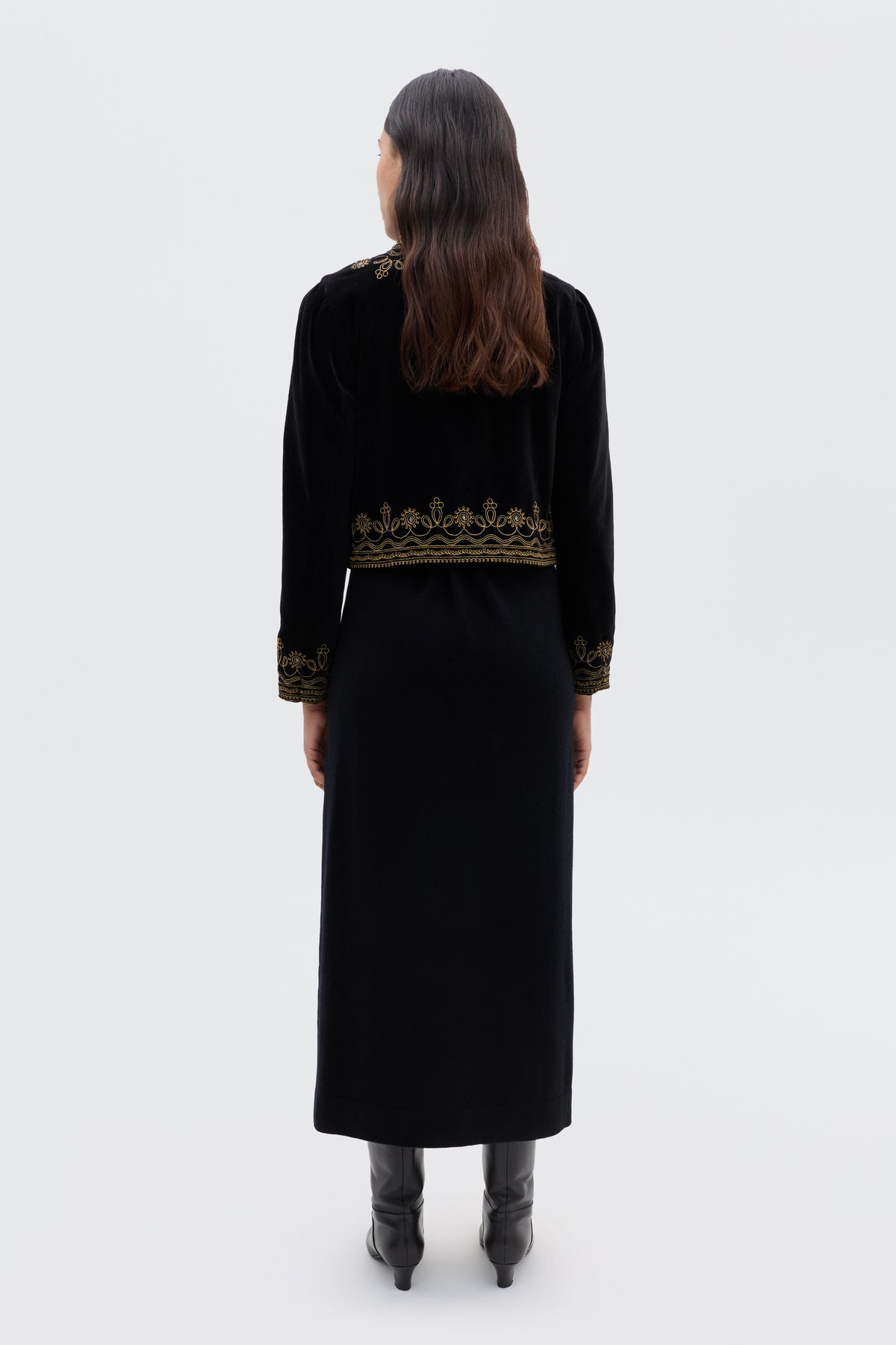A woman with long brown hair, facing away, wears the Denver - Soutache Black: a black velvet jacket with gold embroidery on the hem and cuffs, over a long black dress and boots. The background is plain light gray.