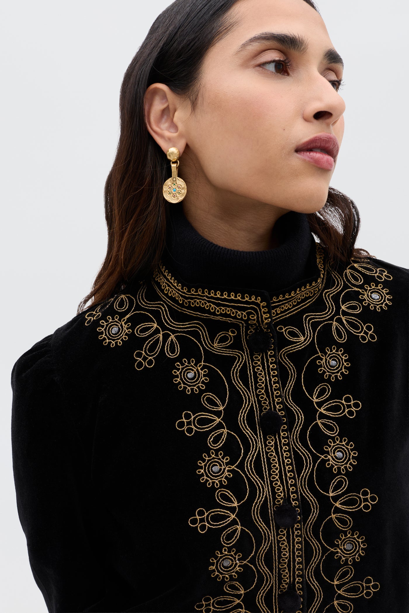 A woman with long dark hair models the Denver - Soutache Black jacket, crafted from cotton-velvet with intricate gold embroidery, paired with gold drop earrings, as she gazes to the side against a plain light background.