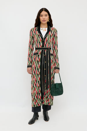 A woman stands against a white background wearing the Apollo - Chevron Yarn Dyed Cream duster coat, featuring green, red, and beige geometric patterns, black pants, black ankle boots, and holding a dark green woven handbag.