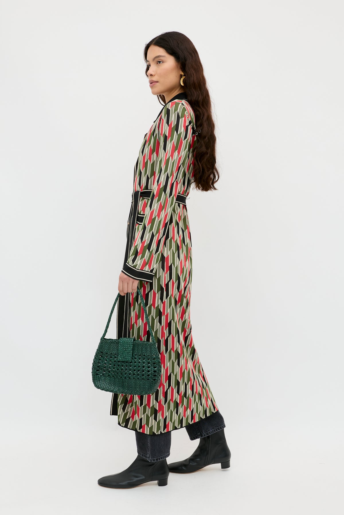 A woman with long dark hair stands sideways against a plain white background, wearing the Apollo - Chevron Yarn Dyed Cream dress, styled with black ankle boots and a green textured handbag.
