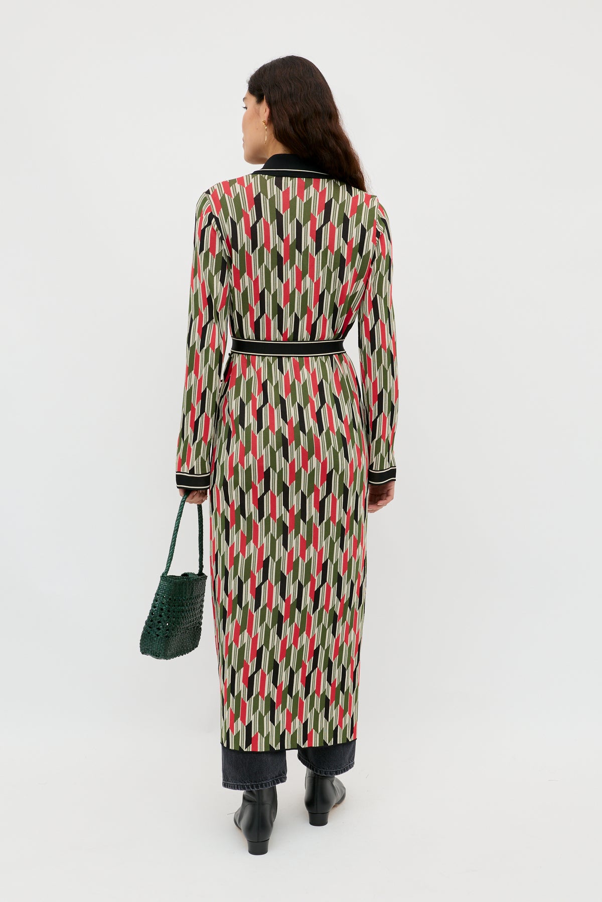 A woman faces away wearing the Apollo - Chevron Yarn Dyed Cream duster with red, black, and beige geometric shapes, paired with black pants and boots. She holds a green textured handbag against a plain white background.
