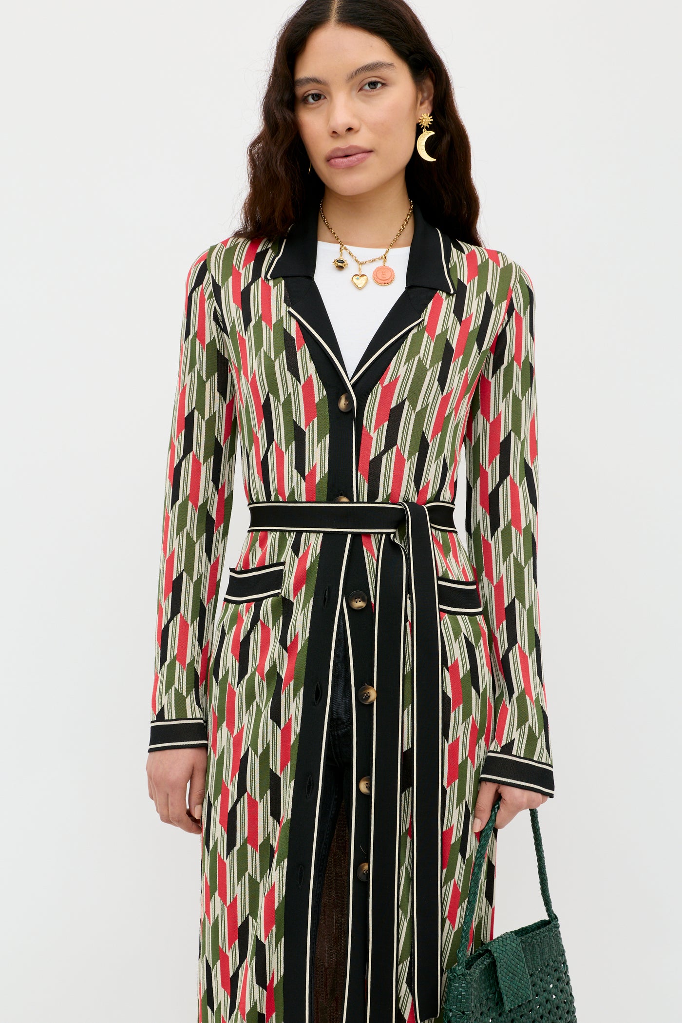 A woman with long brown hair wears the Apollo - Chevron Yarn Dyed Cream coat in a '70s geometric pattern, featuring black lapels, a tie belt, gold buttons over a white top, and carries a green woven handbag.
