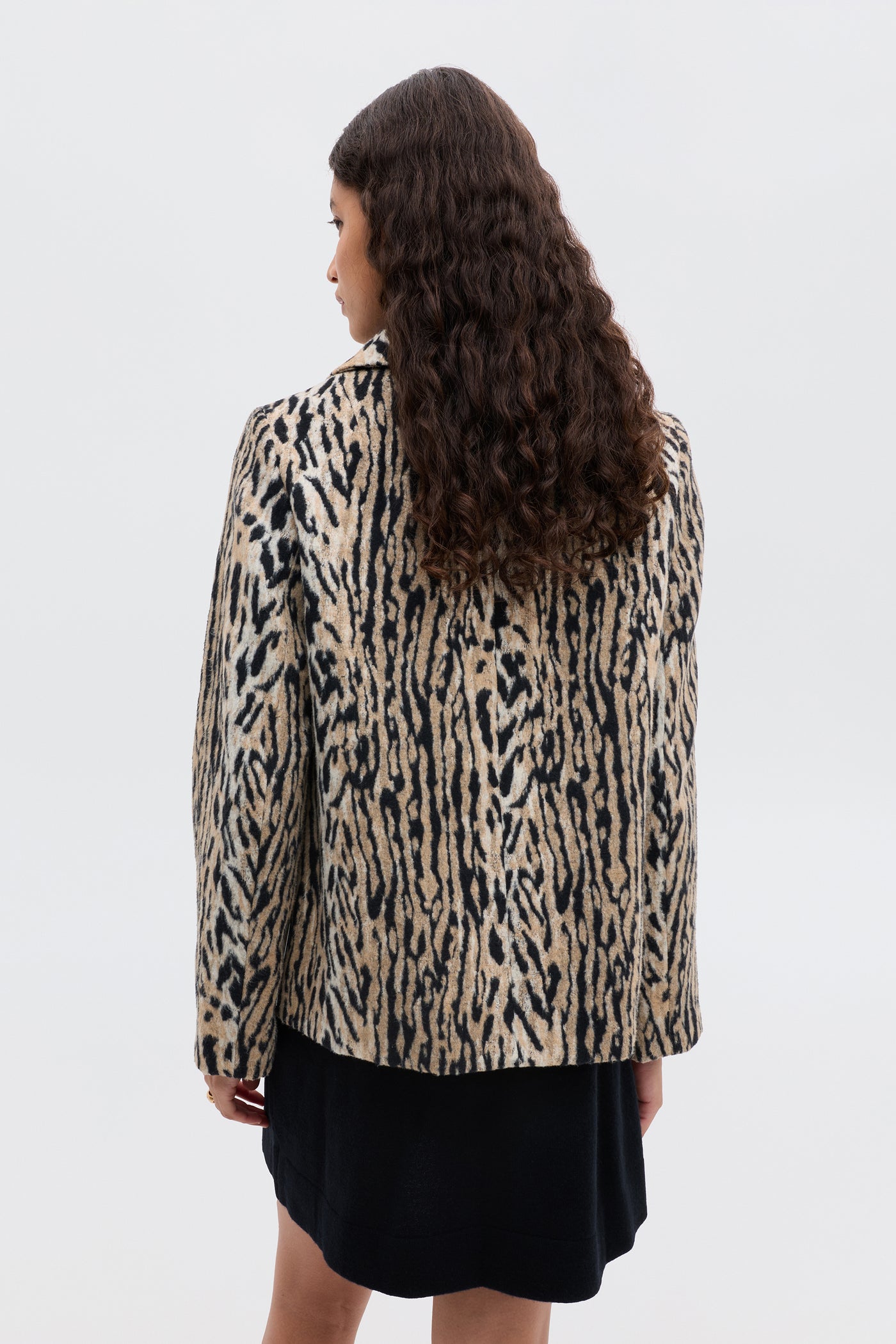 A person with long, curly brown hair stands facing away, wearing the Tommie - Bohemia Leopard jacket over a black dress against a plain light background.