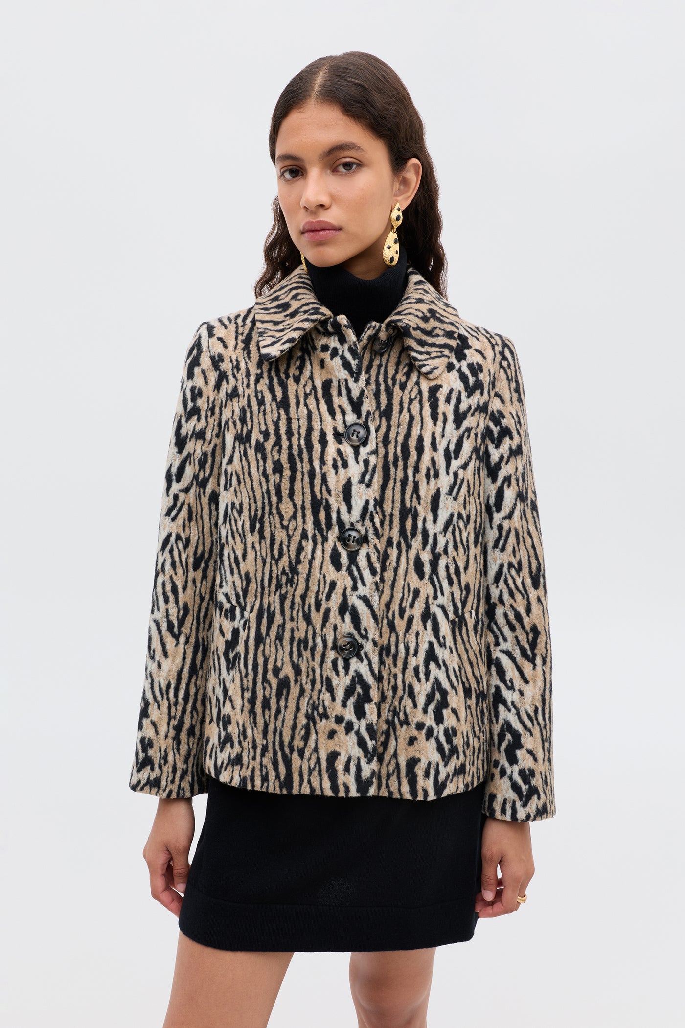A woman poses against a plain background in a black turtleneck mini dress and the Tommie - Bohemia Leopard coat, featuring a beige-and-black animal print with large buttons. Her look is finished with long wavy hair and gold earrings.