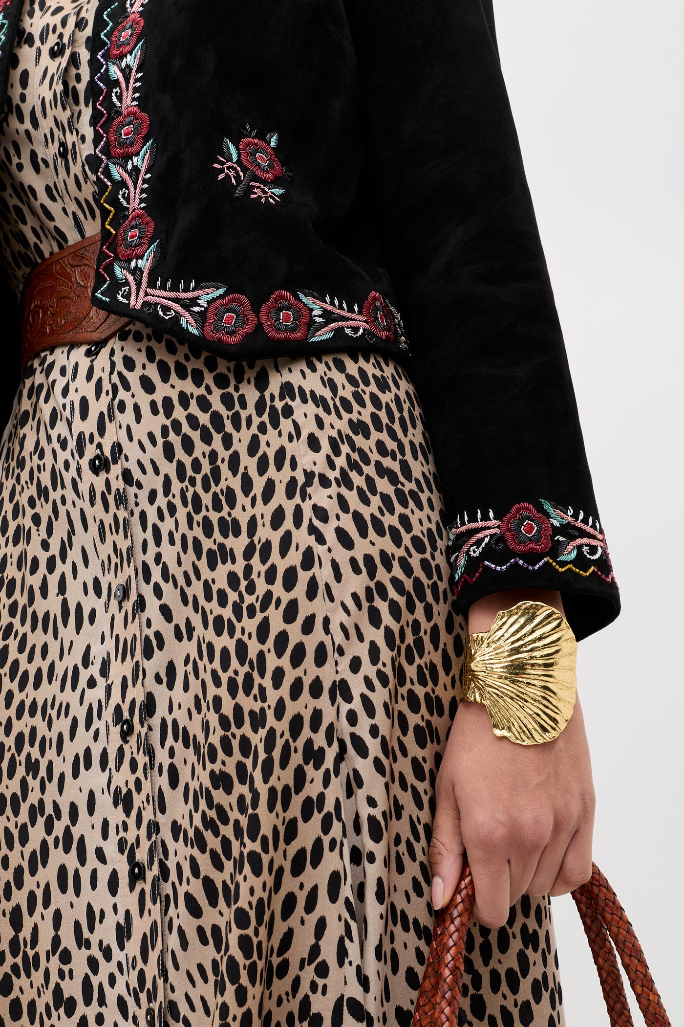 A person wearing the Oliwia - Roselle Embellishment Black jacket, an animal print dress, a brown belt, and a gold shell-shaped bracelet holds a brown woven handle.