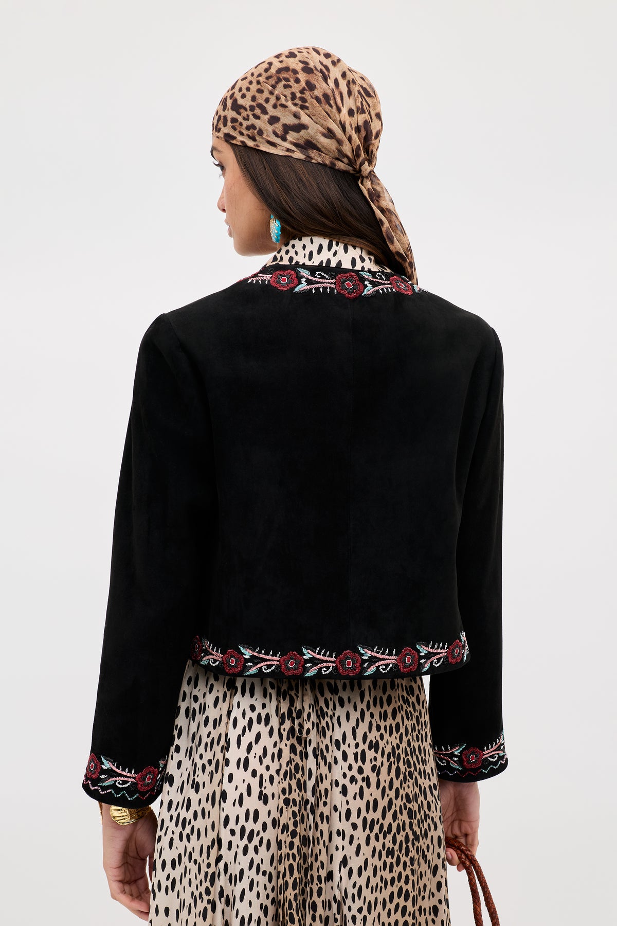 A woman seen from behind wears the Oliwia - Roselle Embellishment Black jacket, paired with a leopard-print headscarf and matching dress. She also accessorizes with turquoise earrings and carries a brown woven bag.
