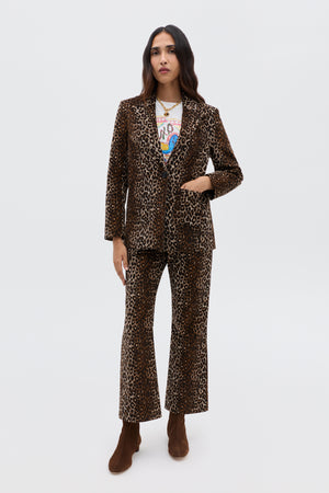 A woman with long, dark hair and a neutral expression wears the Xander - Lyra Leopard Black jacket, paired with flared pants, a graphic tee, brown ankle boots, and layered necklaces, standing against a plain light background.