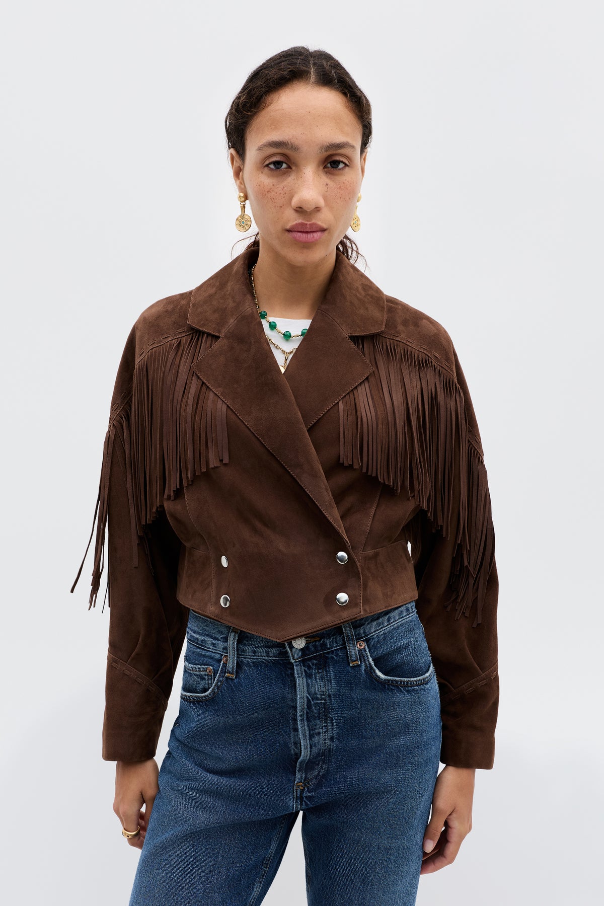 A woman stands against a white background wearing the Darragh - Brown suede jacket with 1970s-inspired fringe detailing, paired with blue high-waisted jeans.