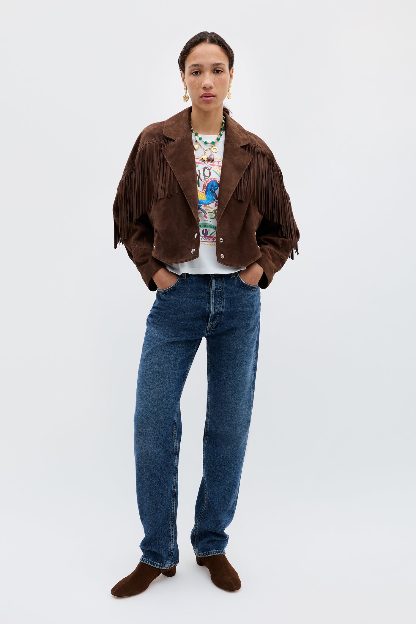 A person stands facing forward, wearing the Darragh - Brown suede jacket over a graphic t-shirt, blue jeans, and brown shoes, hands in pockets against a plain light background.