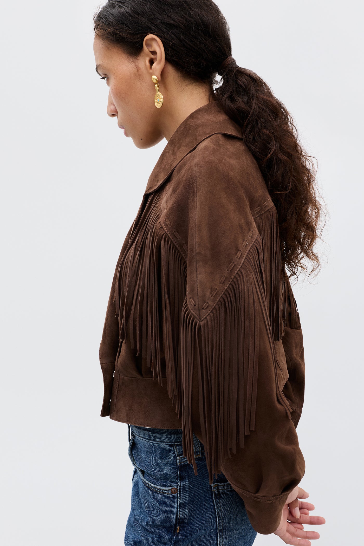 A woman with long curly hair in a ponytail wears gold earrings, blue jeans, and the Darragh - Brown jacket with fringe details, standing in profile against a plain light background.