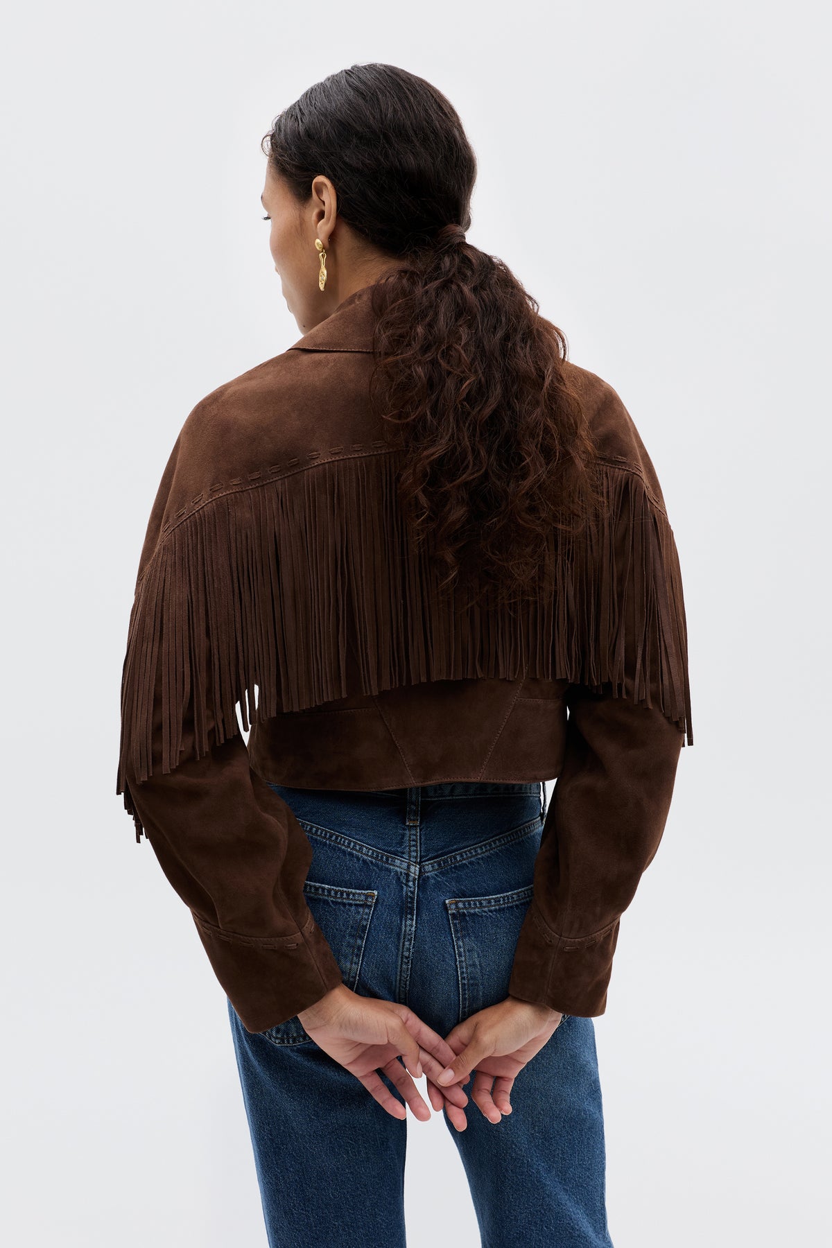 A person with long curly hair in a ponytail, wearing the Darragh - Brown suede jacket with fringe and blue jeans, stands facing away with their hands clasped behind them against a plain background.