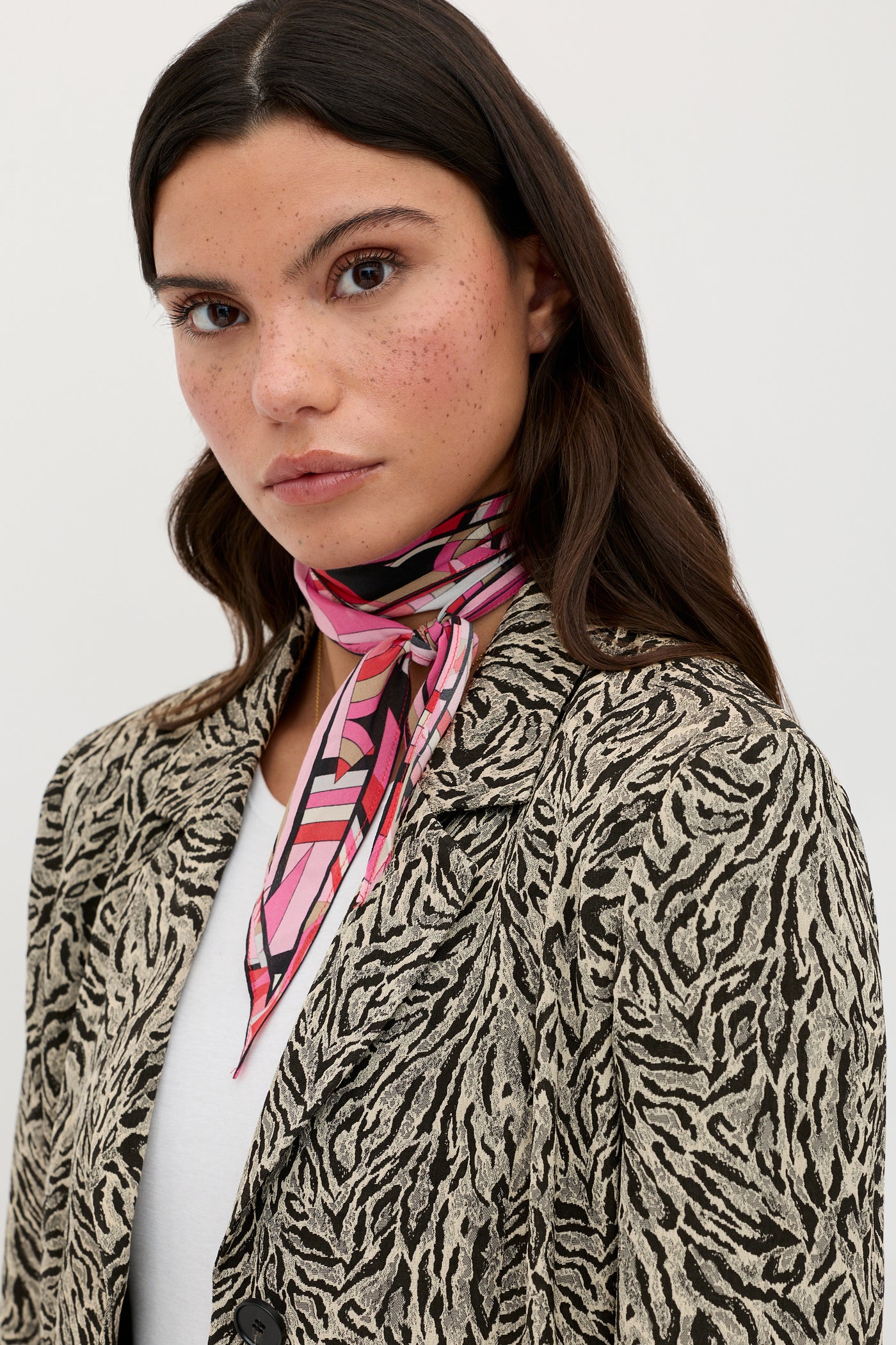 A woman with long dark hair wears the Marselle - Sun Leo Tan patterned beige and black blazer over a white top, accessorized with a pink geometric scarf around her neck. She has a neutral expression and stands against a plain, light background.