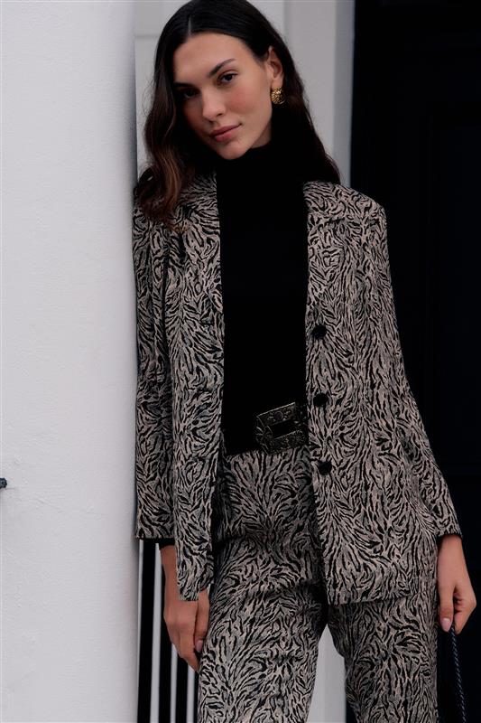 A woman wearing a black turtleneck and a beige patterned suit leans against a white wall, looking at the camera. She has long brown hair, gold earrings, and carries the Marselle - Sun Leo Tan bag by her side.