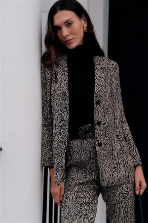 A woman wearing a black turtleneck and a beige patterned suit leans against a white wall, looking at the camera. She has long brown hair, gold earrings, and carries the Marselle - Sun Leo Tan bag by her side.