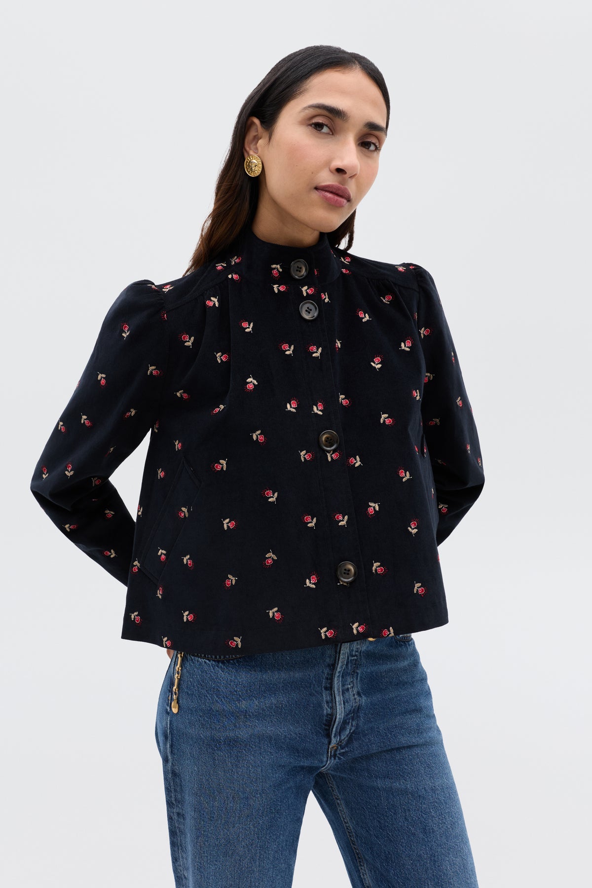 A woman wears the Nyla - Dandy Embroidery Deep Navy jacket, featuring a '60s-inspired cropped fit and small floral print, styled with gold button earrings and blue jeans, standing against a plain light background with a slight smile.