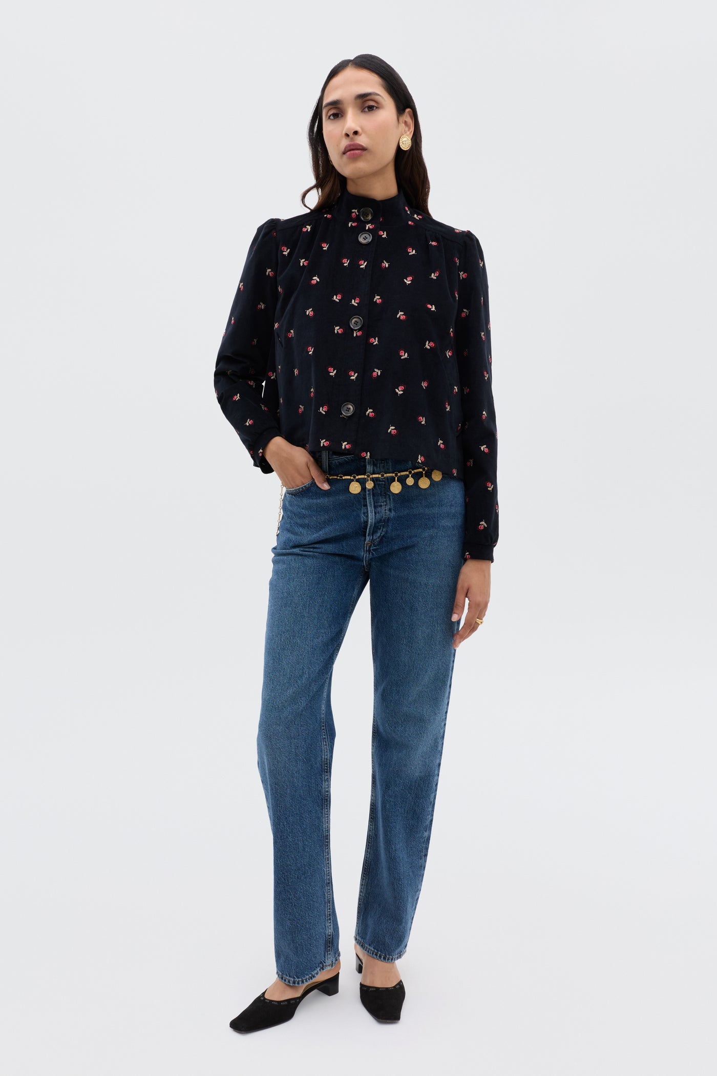 A woman stands against a plain background wearing the Nyla - Dandy Embroidery Deep Navy blouse by RIXO, featuring long sleeves and a floral pattern, paired with blue jeans, black shoes, and a decorative belt with round gold accents.