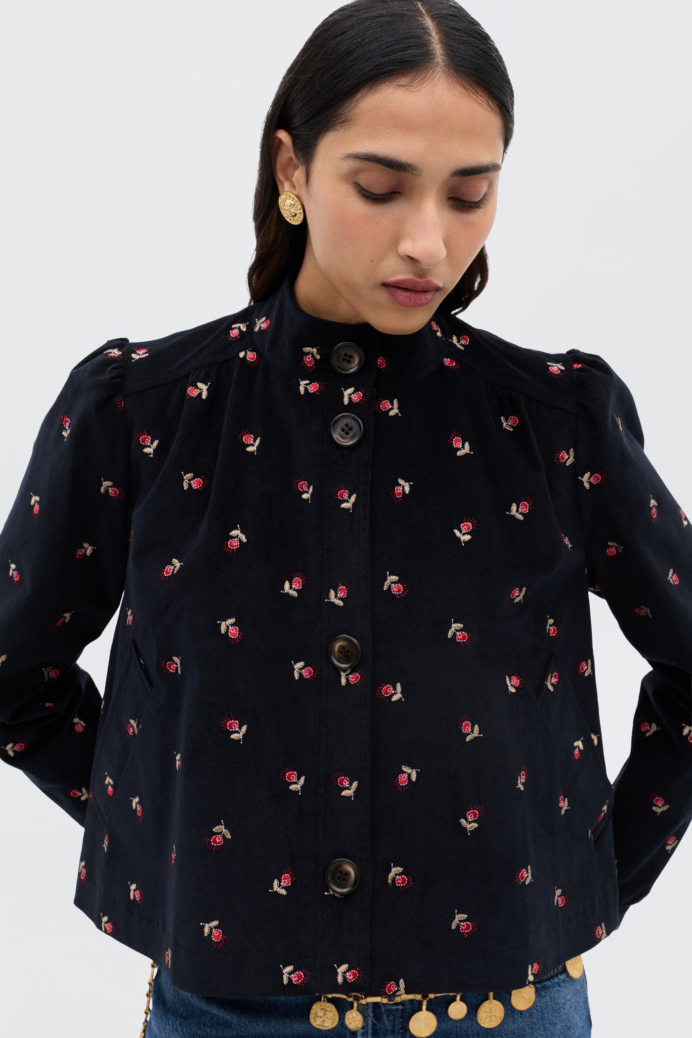 A person wearing the Nyla - Dandy Embroidery Deep Navy jacket with small red and beige floral details, gold button earrings, and blue jeans looks downward against a plain light background.