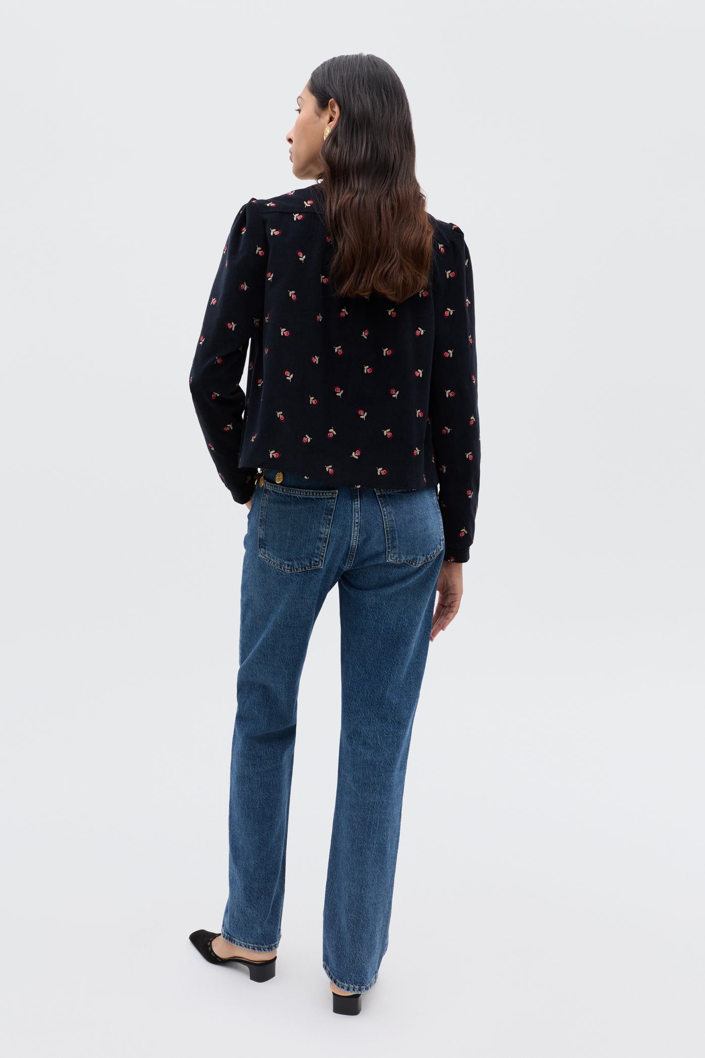 A woman with long brown hair, facing away, wears the Nyla - Dandy Embroidery Deep Navy top with small red floral patterns, cropped blue straight-leg jeans, and black flats against a plain light background.