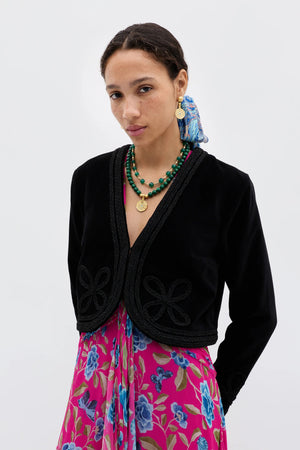 A woman with dark braided hair, gold earrings, and layered green necklaces models the Kesia - Clover Appliqué Black, a vintage-inspired velvet jacket with decorative trim, over a bright pink floral dress against a white background.