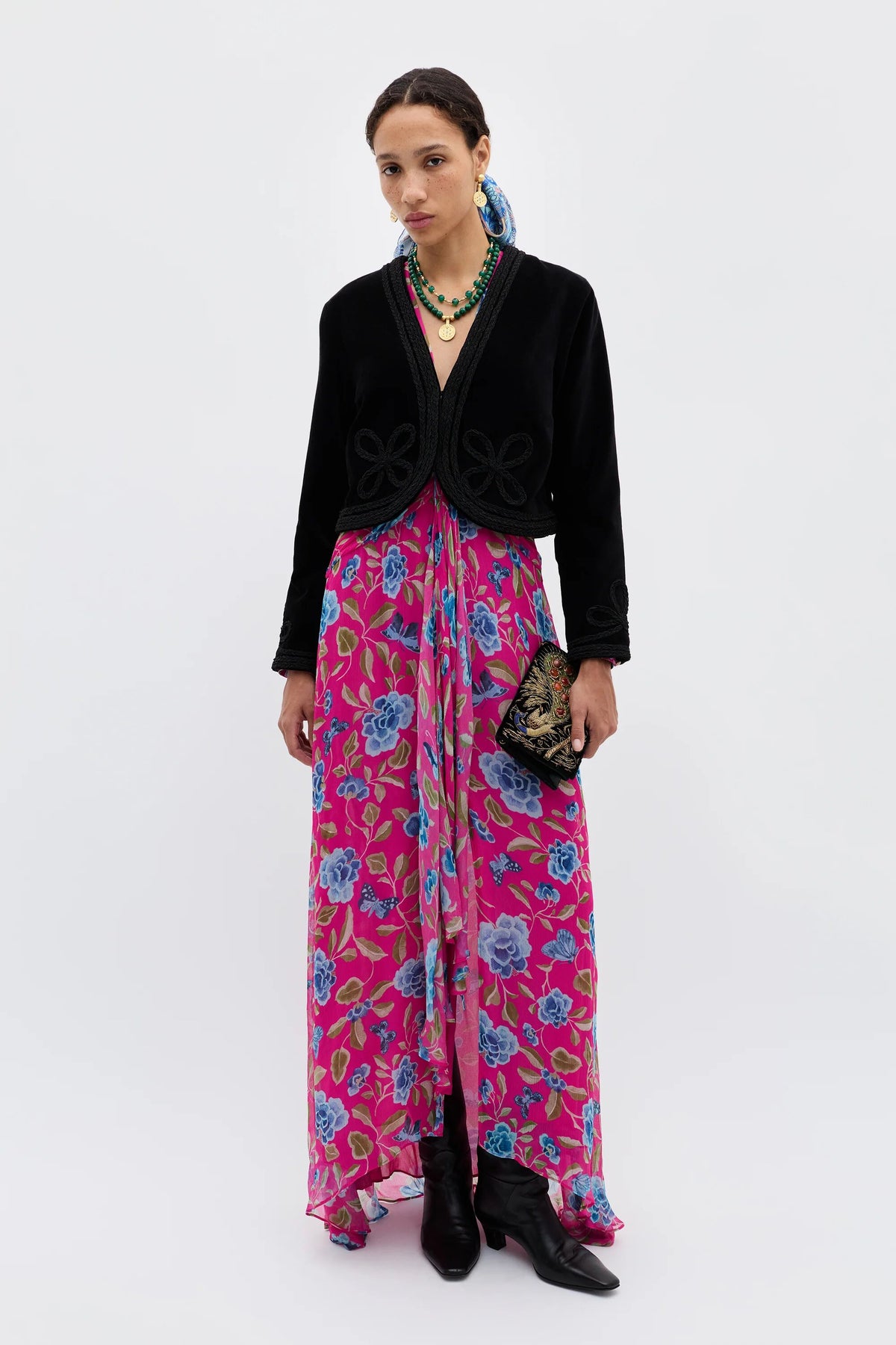 A woman stands against a plain background wearing the Kesia - Clover Appliqué Black jacket over a bright pink floral dress, paired with black boots, layered necklaces, and her hair styled back as she holds a decorative clutch.