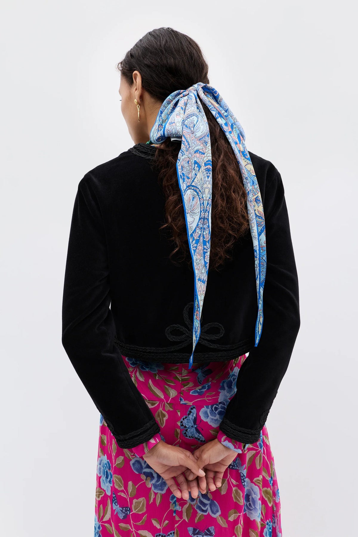 Seen from behind, a person with long dark hair wears the Kesia - Clover Appliqué Black jacket and a vibrant blue and white scarf in their hair, paired with a pink dress featuring blue floral patterns, hands gently clasped behind their back.