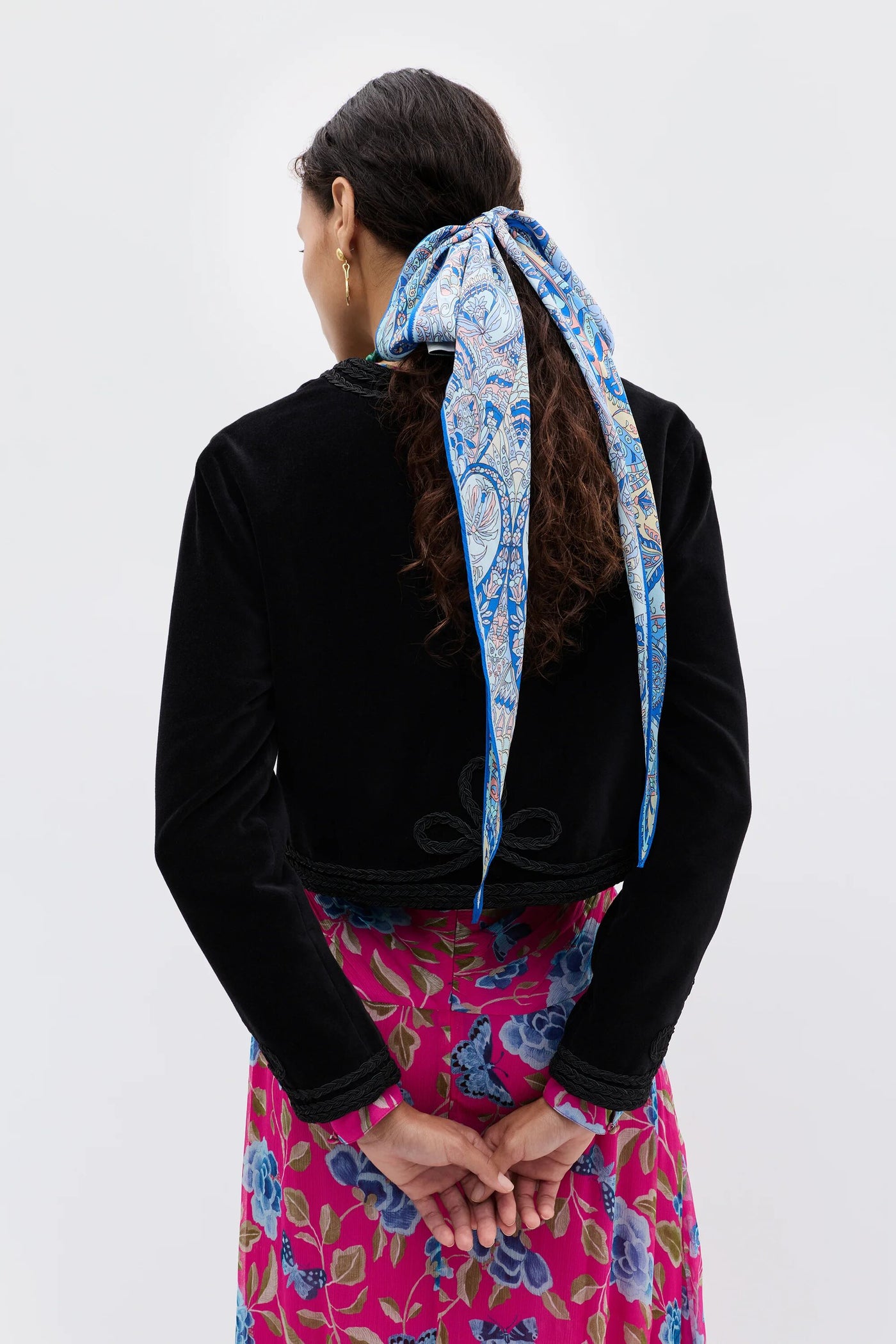Seen from behind, a person with long dark hair wears the Kesia - Clover Appliqué Black jacket and a vibrant blue and white scarf in their hair, paired with a pink dress featuring blue floral patterns, hands gently clasped behind their back.
