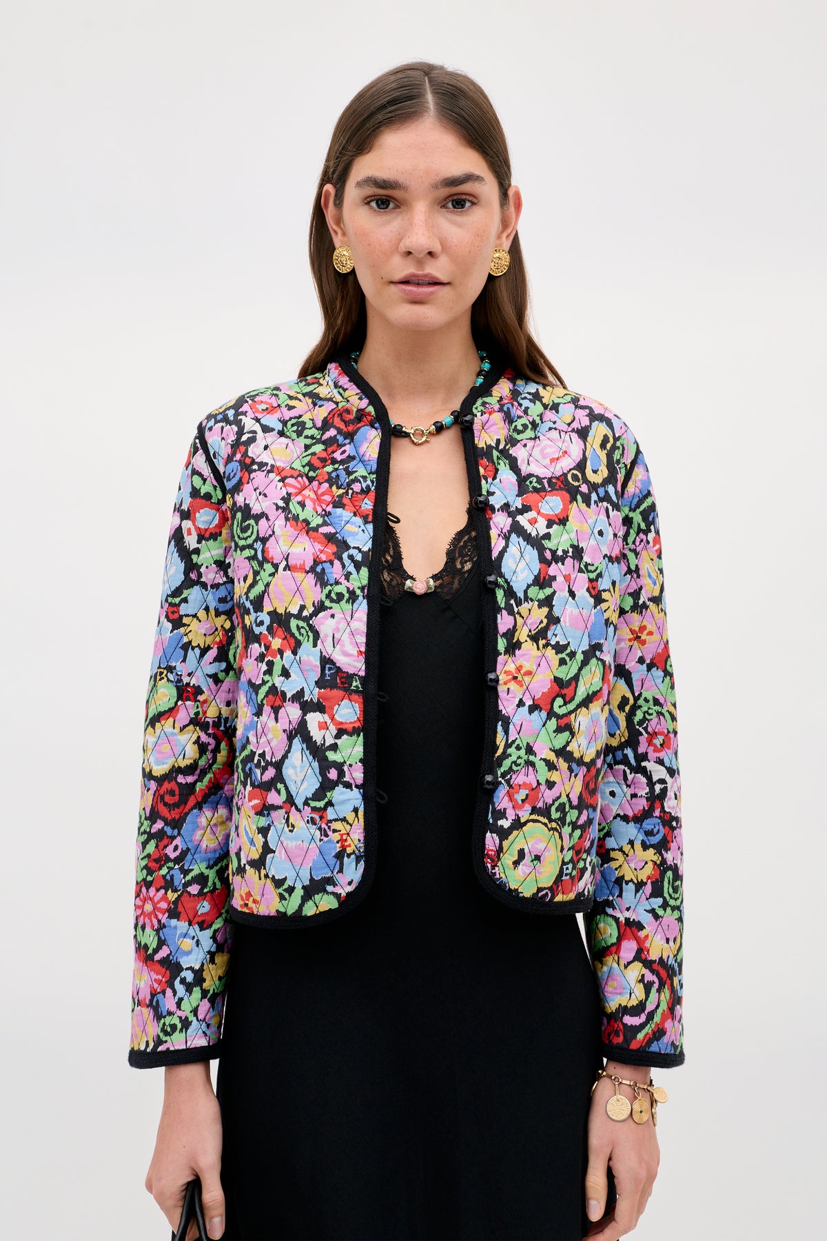 A woman wears the Lyanne - Ikat Bloom Charcoal jacket, featuring a colorful floral pattern over a black dress, accessorized with gold earrings, a necklace, and bracelets, standing against a plain light background.
