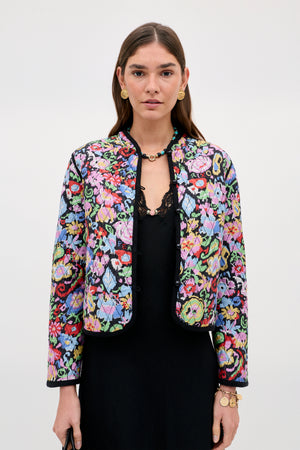 A woman wears the Lyanne - Ikat Bloom Charcoal jacket, featuring a colorful floral pattern over a black dress, accessorized with gold earrings, a necklace, and bracelets, standing against a plain light background.