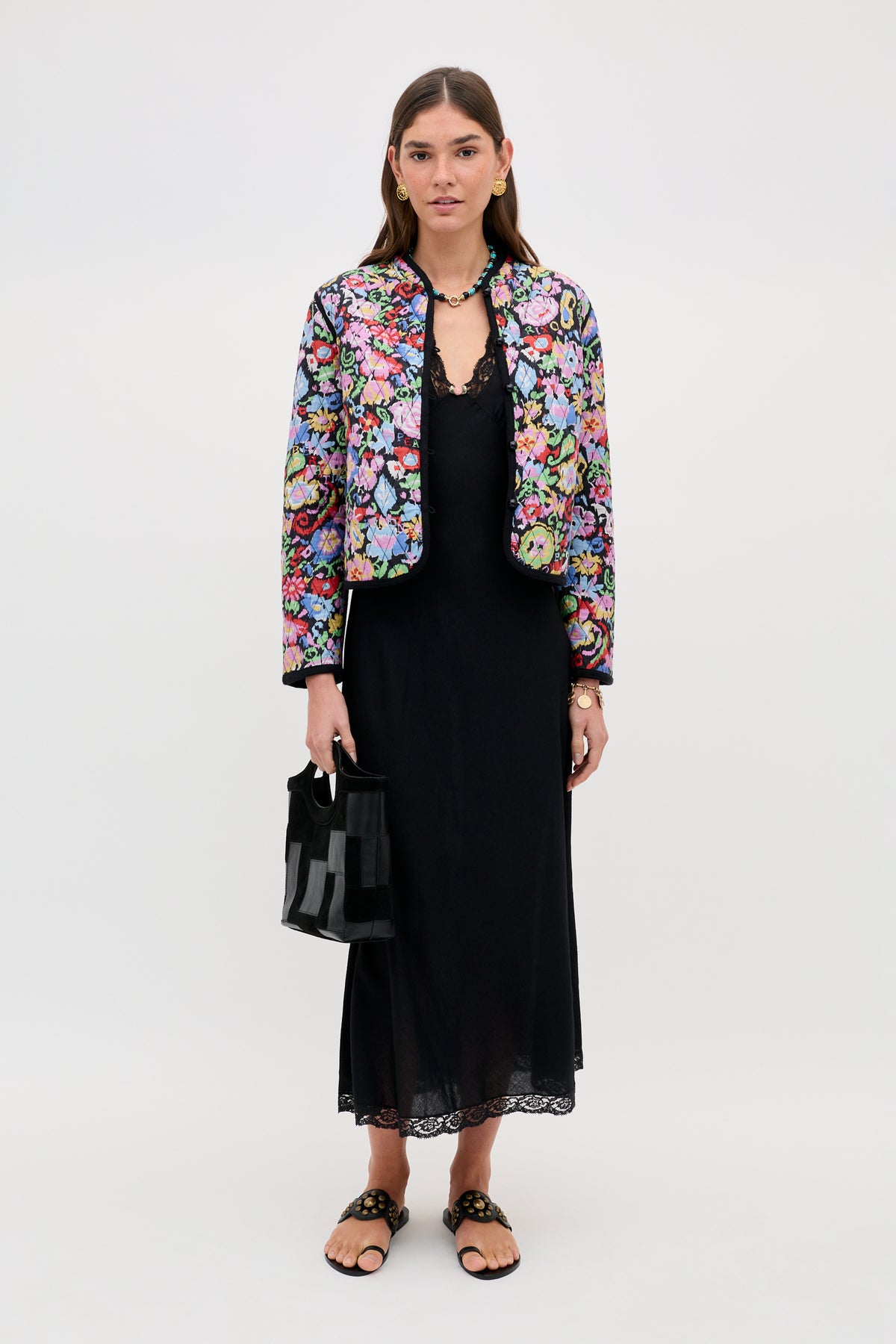 A woman with long brown hair and minimal makeup stands against a plain background wearing the Lyanne - Ikat Bloom Charcoal jacket over a long black dress, paired with sandals and holding a small black bag.