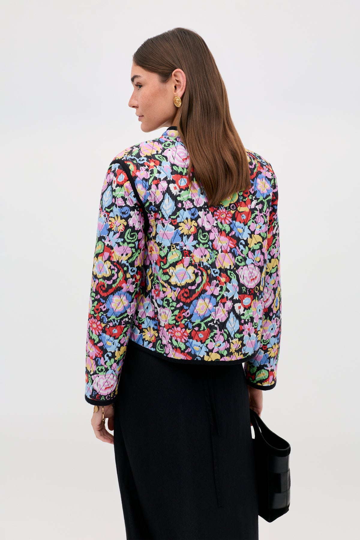 A woman stands with her back to the camera, wearing the Lyanne - Ikat Bloom Charcoal jacket—a vibrant floral-patterned piece—paired with a long black skirt and carrying a small black handbag. Her brown hair is tucked behind her ear.