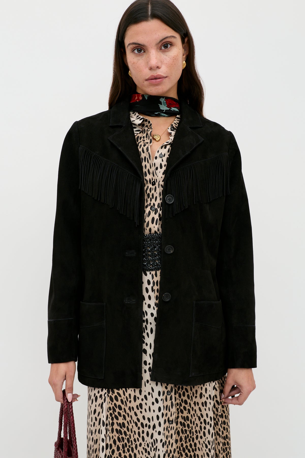 A woman stands against a plain background wearing the Teagan - Black suede jacket with fringe detailing over a leopard print dress, accessorized with a red floral scarf and holding a small textured bag.
