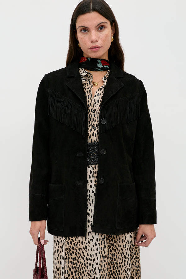 A woman stands against a plain background wearing the Teagan - Black suede jacket with fringe detailing over a leopard print dress, accessorized with a red floral scarf and holding a small textured bag.