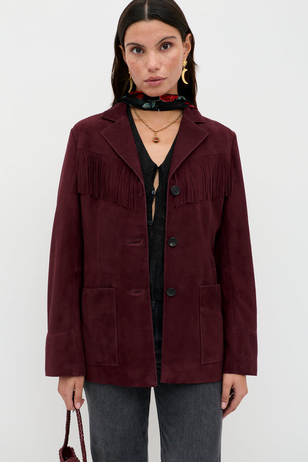 A woman wears the Teagan - Maroon suede fringe jacket, paired with a black shirt, dark jeans, layered necklaces, gold hoops, a floral scarf, and holds a small bag against a plain background.
