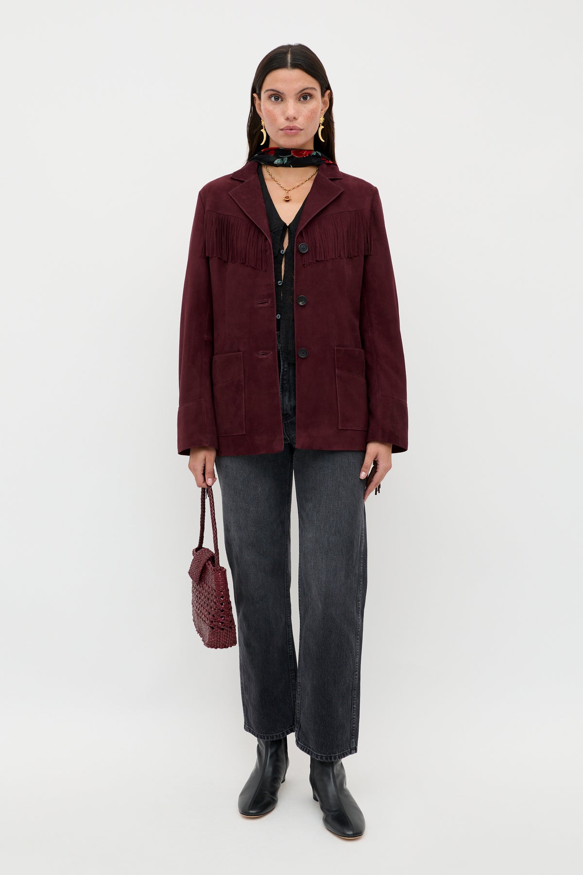 A woman with straight dark hair and layered necklaces stands against a plain white background, wearing the Teagan - Maroon fringe jacket, a black shirt, dark gray jeans, black ankle boots, and holding a red textured handbag.
