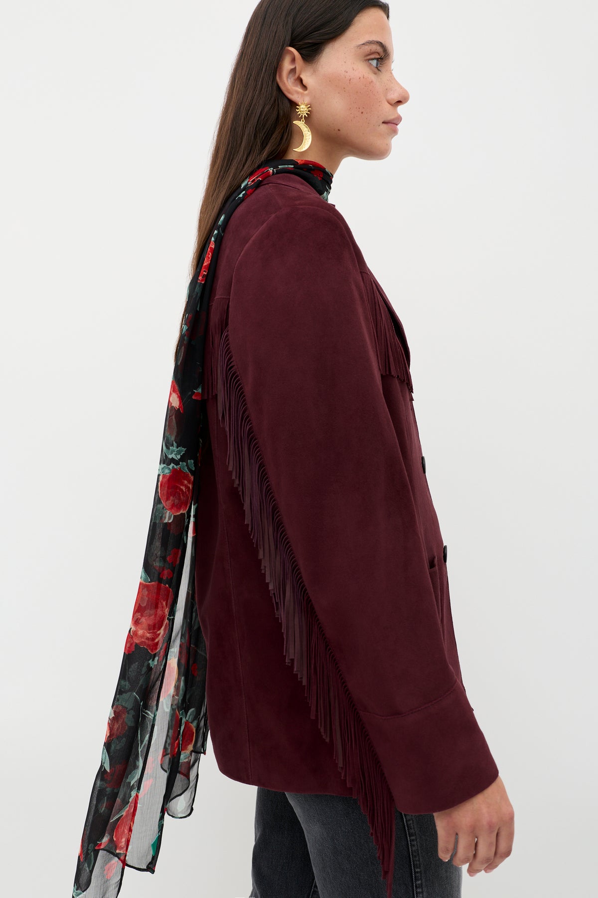 A woman in profile wears the Teagan – Maroon fringed jacket with dark jeans, gold earrings, and a long sheer black scarf with red floral designs. Her straight brown hair is worn down.