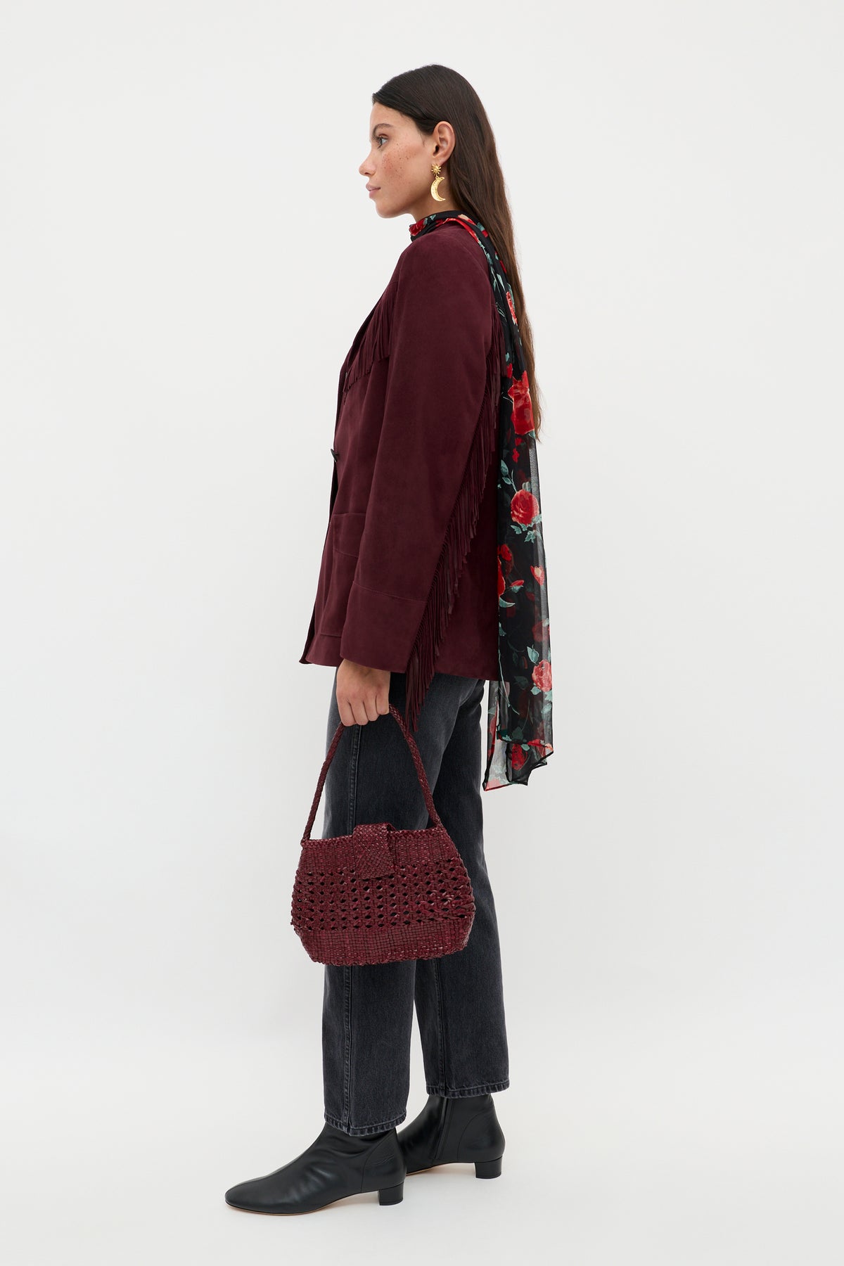 A woman with long dark hair stands in profile against a white background, holding the Teagan - Maroon textured handbag. She wears a maroon fringed jacket, floral scarf, dark jeans, and black ankle boots.