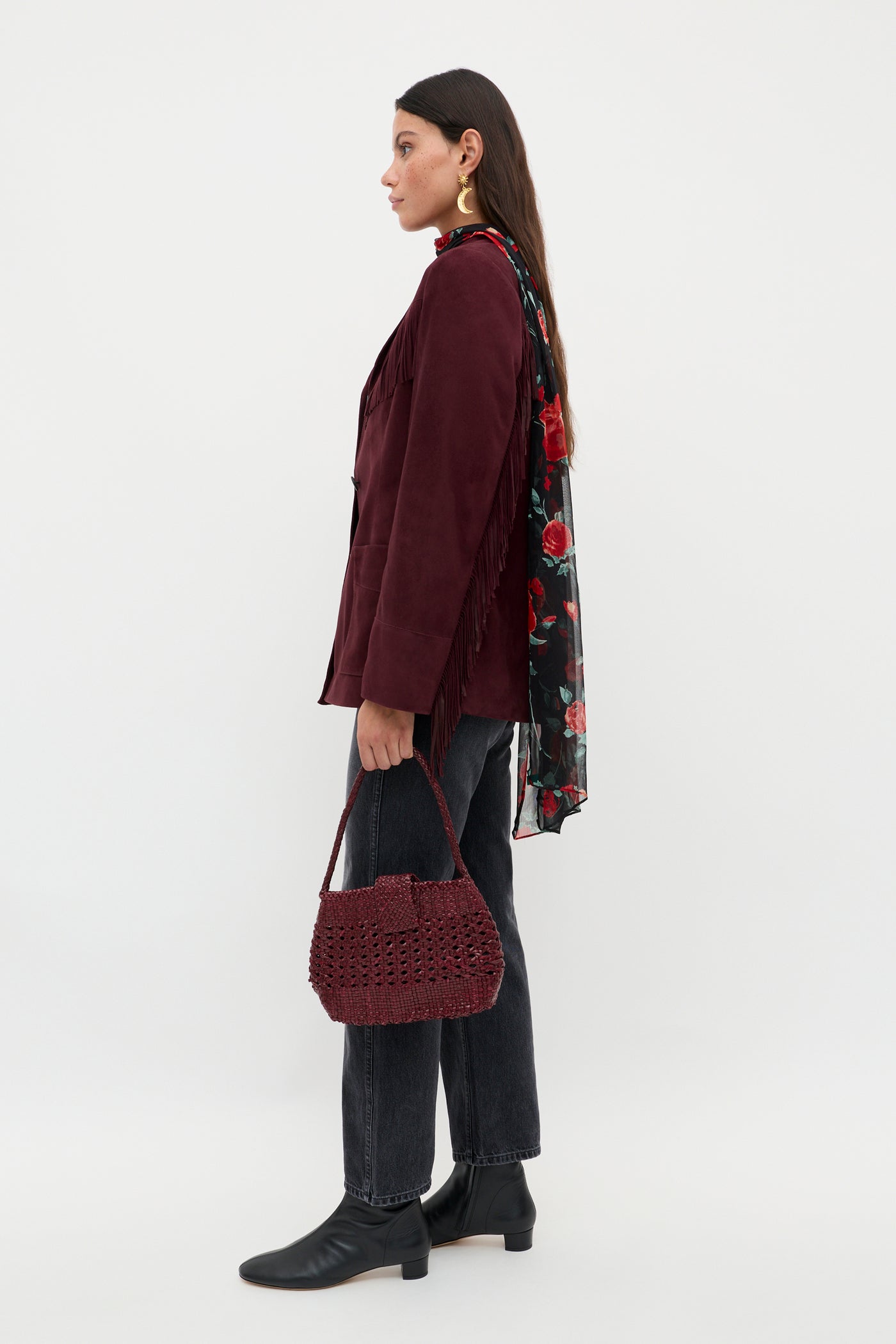 A woman with long dark hair stands in profile against a white background, holding the Teagan - Maroon textured handbag. She wears a maroon fringed jacket, floral scarf, dark jeans, and black ankle boots.