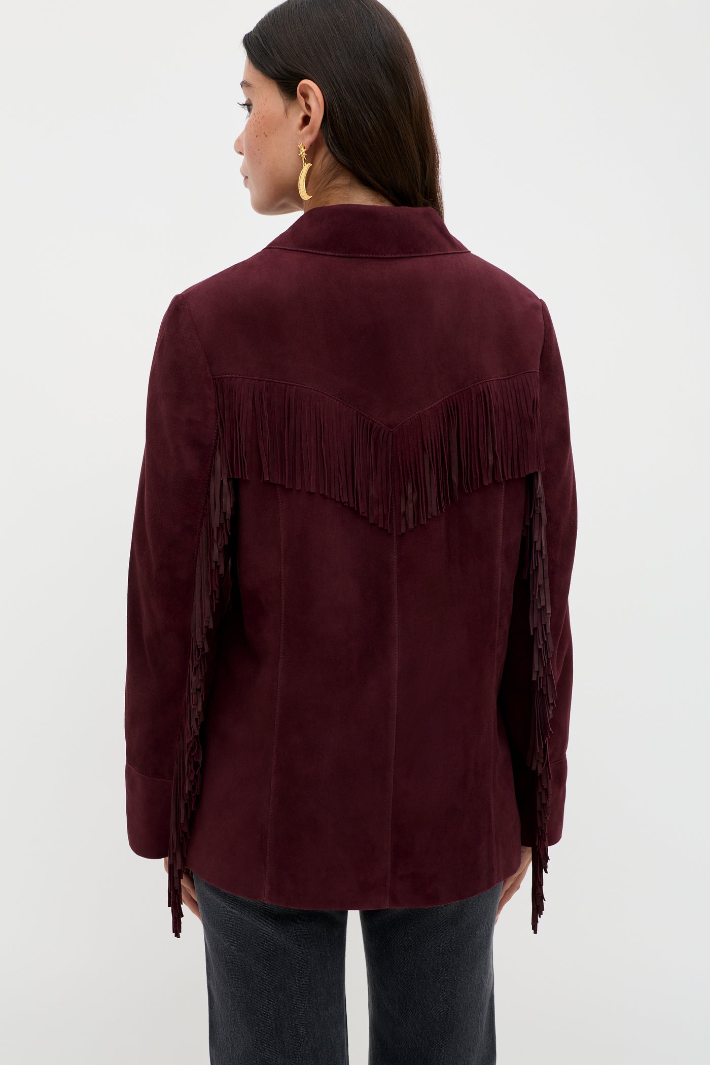 A woman with long dark hair wears the Teagan - Maroon suede jacket, featuring fringe across the back and sleeves. She faces away from the camera, accessorized with gold hoop earrings, against a plain light background.