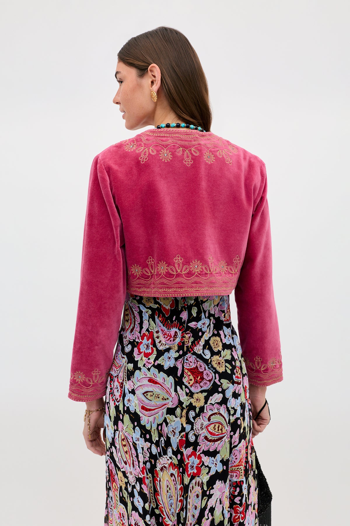 A woman faces away, wearing the Cliona - Soutache Embroidery Magenta Pink velvet jacket with gold detailing and a long, colorful paisley skirt. The backdrop is plain and light-colored.