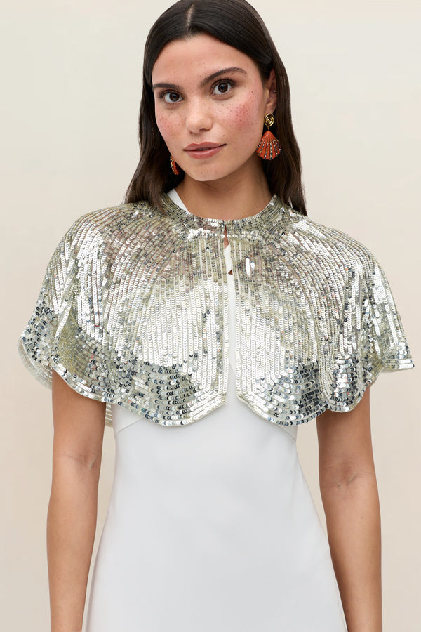 A woman wearing the Evienne Art Deco Embellishment Pearl cape over a white dress looks at the camera. She has long brown hair, natural makeup, and is accessorized with red fan-shaped earrings.