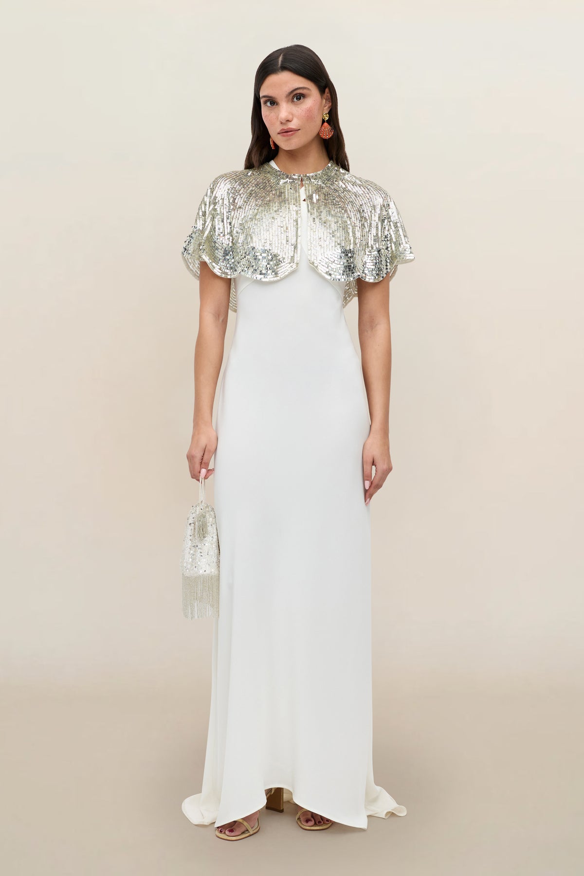 Wearing the Evienne - Art Deco Embellishment Pearl, a woman stands against a neutral background in a floor-length white gown with a shimmering Art Deco cape draped over her shoulders, holding the matching beaded bag—an elegant party accessory.