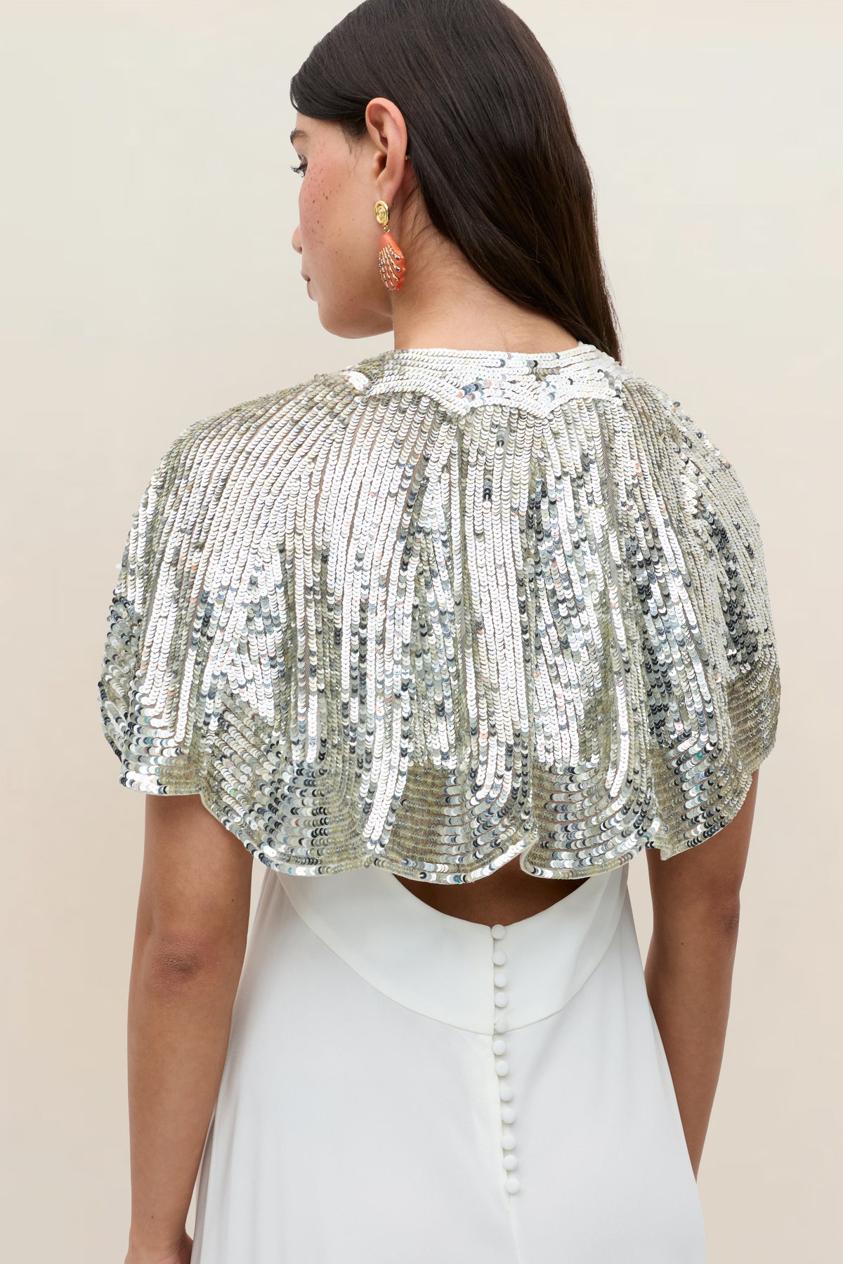 A woman with long dark hair and coral earrings is pictured from the back against a neutral background, wearing a white dress with button details paired with the elegant Evienne – Art Deco Embellishment Pearl cape, which shimmers as a standout party accessory.