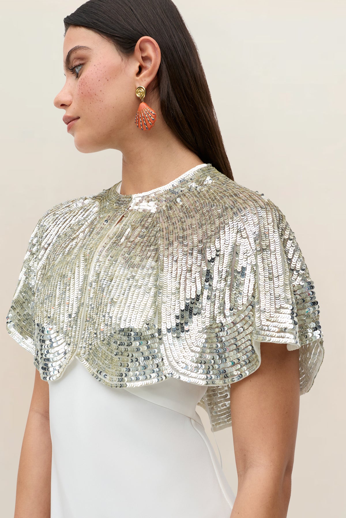Wearing the “Evienne - Art Deco Embellishment Pearl,” a woman in a white dress showcases this sparkling, silver sequin capelet inspired by classic Art Deco style. With long brown hair and gold and red earrings, she poses in profile against a plain beige background—a dazzling party accessory perfect for any festive occasion.