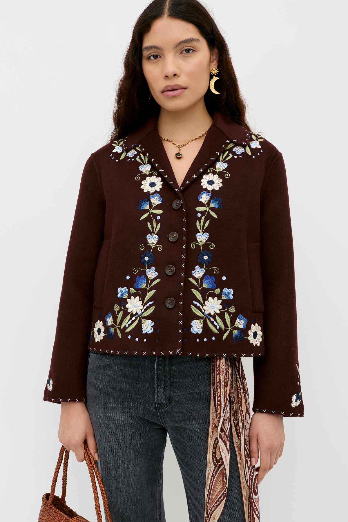 A woman with long dark hair wears the Jude - Bliss Embroidery Brown jacket, styled with black jeans, gold jewelry, a brown woven bag, and a patterned scarf against a plain white background.