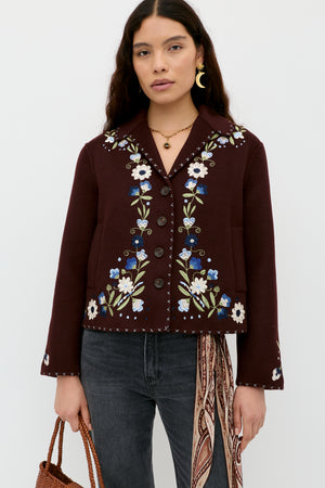 A woman with long dark hair wears the Jude - Bliss Embroidery Brown jacket, styled with black jeans, gold jewelry, a brown woven bag, and a patterned scarf against a plain white background.