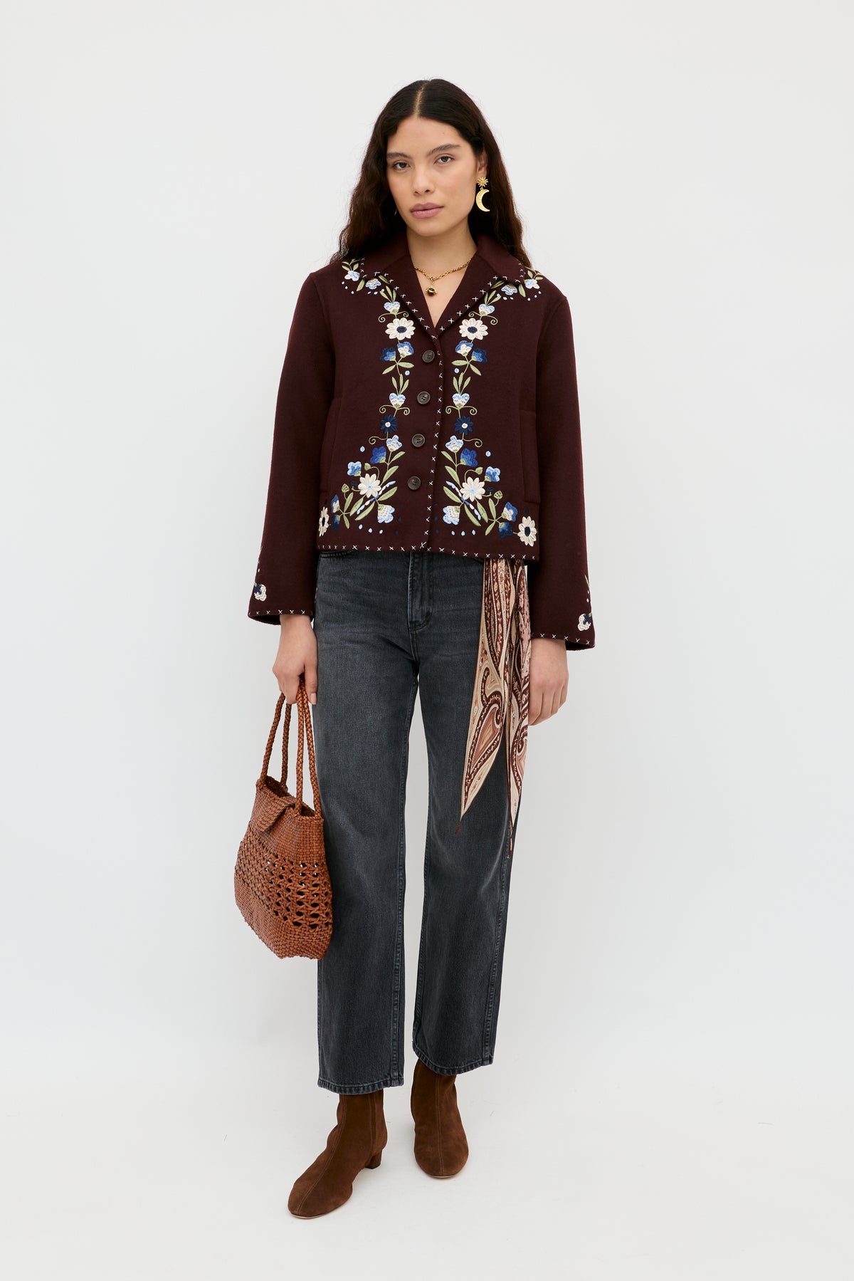 A woman with long dark hair stands against a white background wearing the Jude - Bliss Embroidery Brown jacket by RIXO, dark jeans, brown ankle boots, a patterned scarf tucked in her jeans, and holding a brown woven bag.